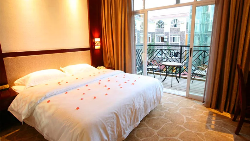 Kunming Tangyun Hotel 昆明飯店客房內部景觀,一張鋪著白色床單的大床上點綴玫瑰花瓣,營造溫馨浪漫氛圍。房間採暖色燈光與木質裝潢設計,落地玻璃門通往陽台,陽台擺放桌椅並可看到外部建築景色,展現昆明酒店舒適與度假感。