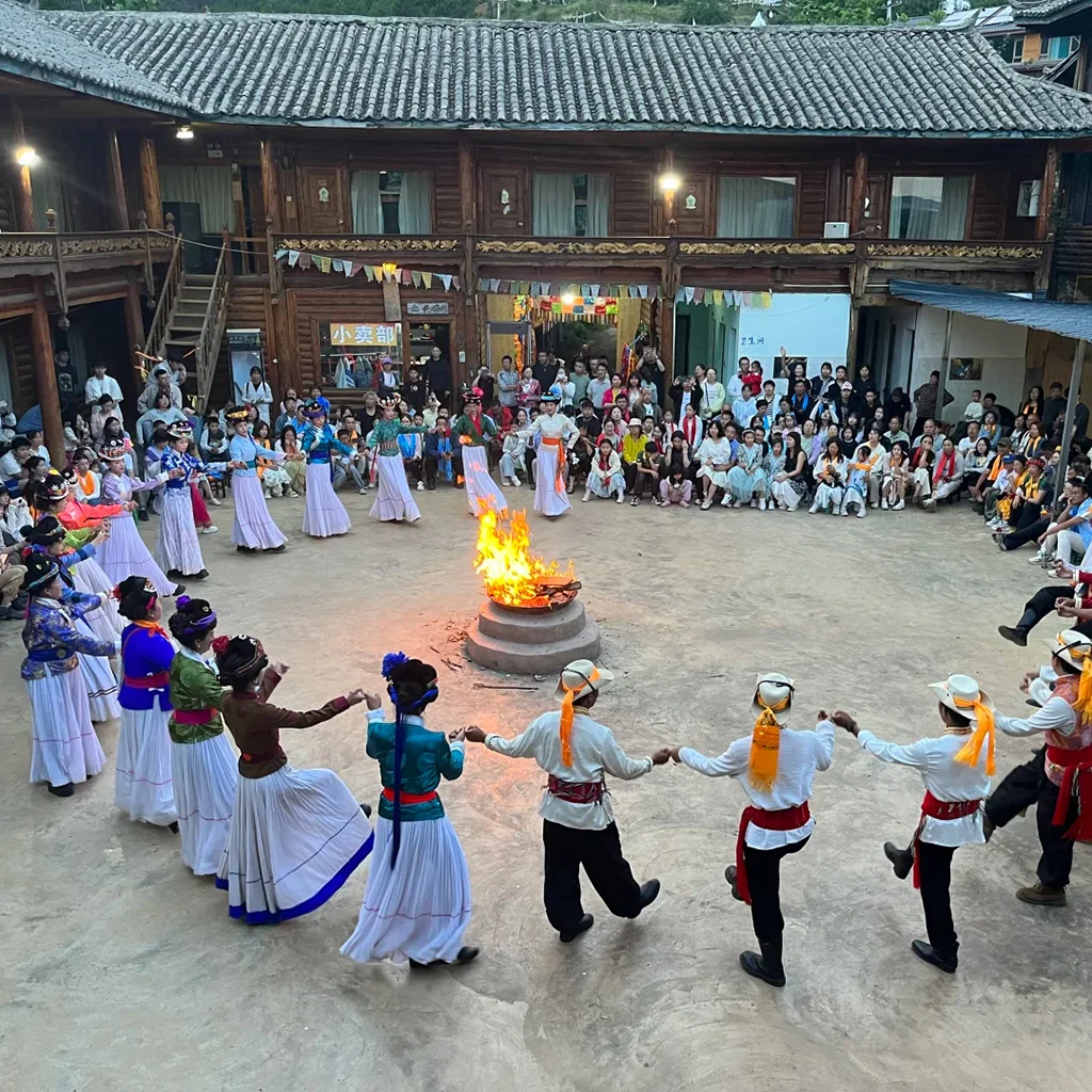 Mosuo Bonfire Party 瀘沽湖篝火晚會。身穿傳統摩梭服飾的男女手拉手圍成一圈,繞著巨大的點燃火坑起舞,身後是落座的觀眾和一座多層木建築。