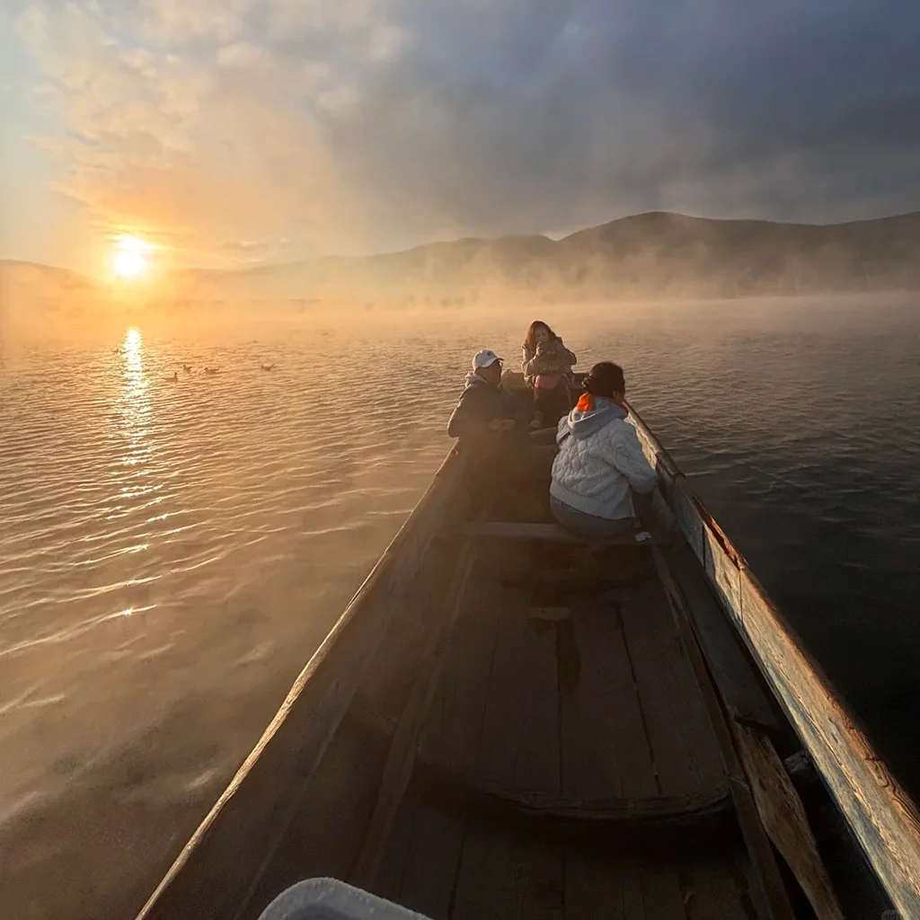 pig trough boats on Lugu Lake 2 雲南瀘沽湖的寧靜日出船遊。傳統木製豬槽船載著遊客,滑行在霧氣瀰漫的湖面,背景是金燦燦的日出與山景。