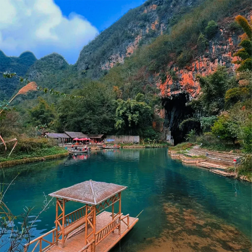 Bamei Cave 位於 坝美溶洞 的湖泊與溶洞入口景觀。清澈湖水倒映群山與森林,岸邊設有竹木亭與步道,遠處紅色岩壁下的巨大溶洞口通向山體內部。