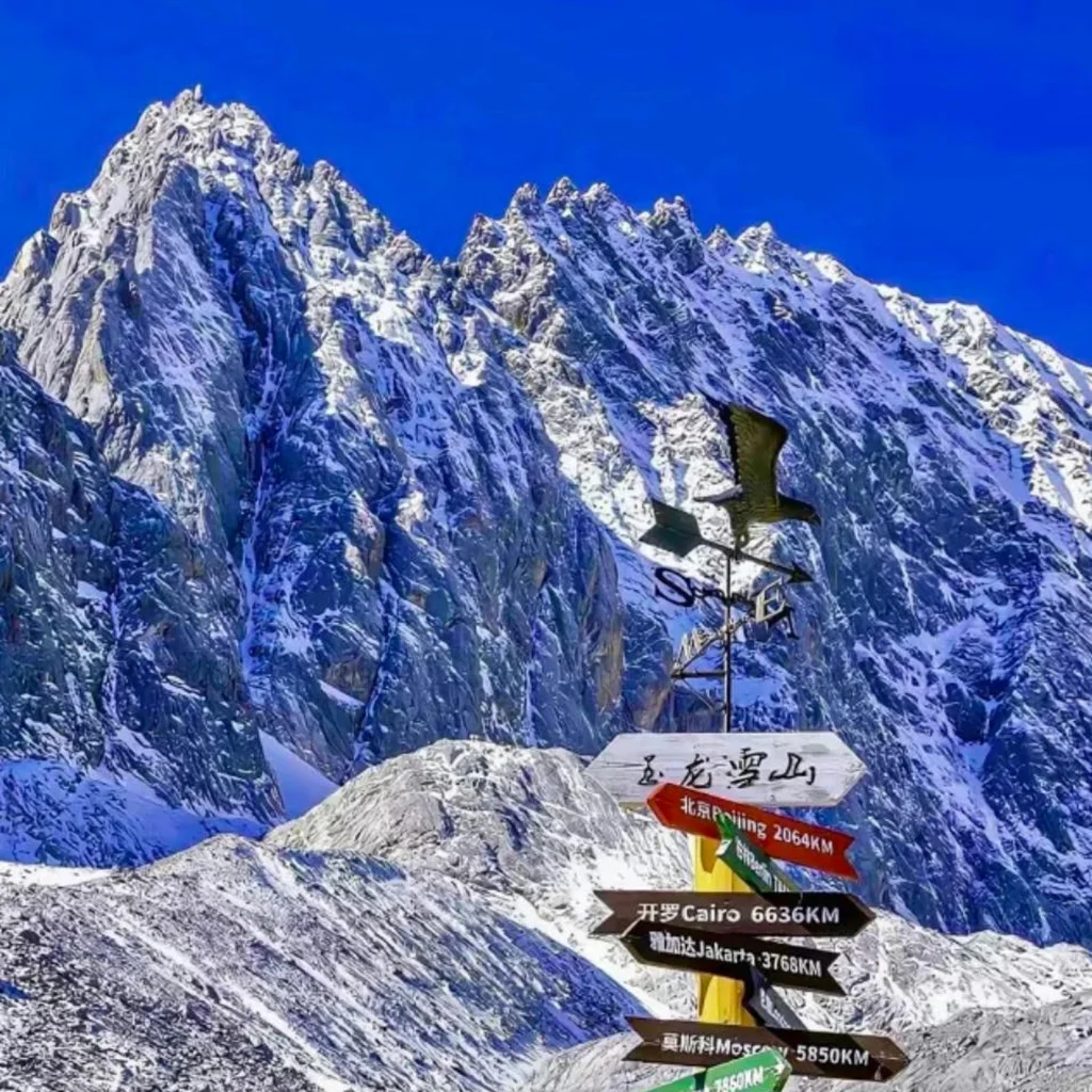Jade Dragon Snow Mountain 高聳陡峭的雪山覆蓋著白雪,在湛藍天空下顯得格外壯觀。前景是一個寫著「玉龍雪山」的方向指示牌與風向標,指向世界多個城市距離,為畫面增添旅遊地標特色,展現出這座著名雪山的雄偉與魅力。