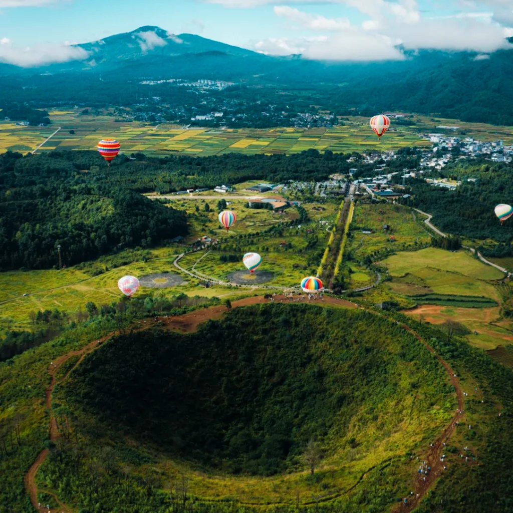 Tengchong Volcanic Geothermal National Geopark 騰沖火山地熱國家地質公園壯觀火山口地形與熱氣球景觀,從高空俯瞰完整火山錐與綠色森林環繞,多彩熱氣球緩緩升空,遠處田園與山巒相連,展現騰沖火山地熱國家地質公園獨特火山地貌與自然生態景觀。