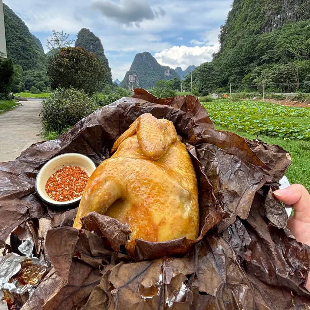 He Ye Chicken 一整隻放在乾燥荷葉上的烤雞,旁附辣椒香料,背景為普者黑標誌性的喀斯特山脈景觀。